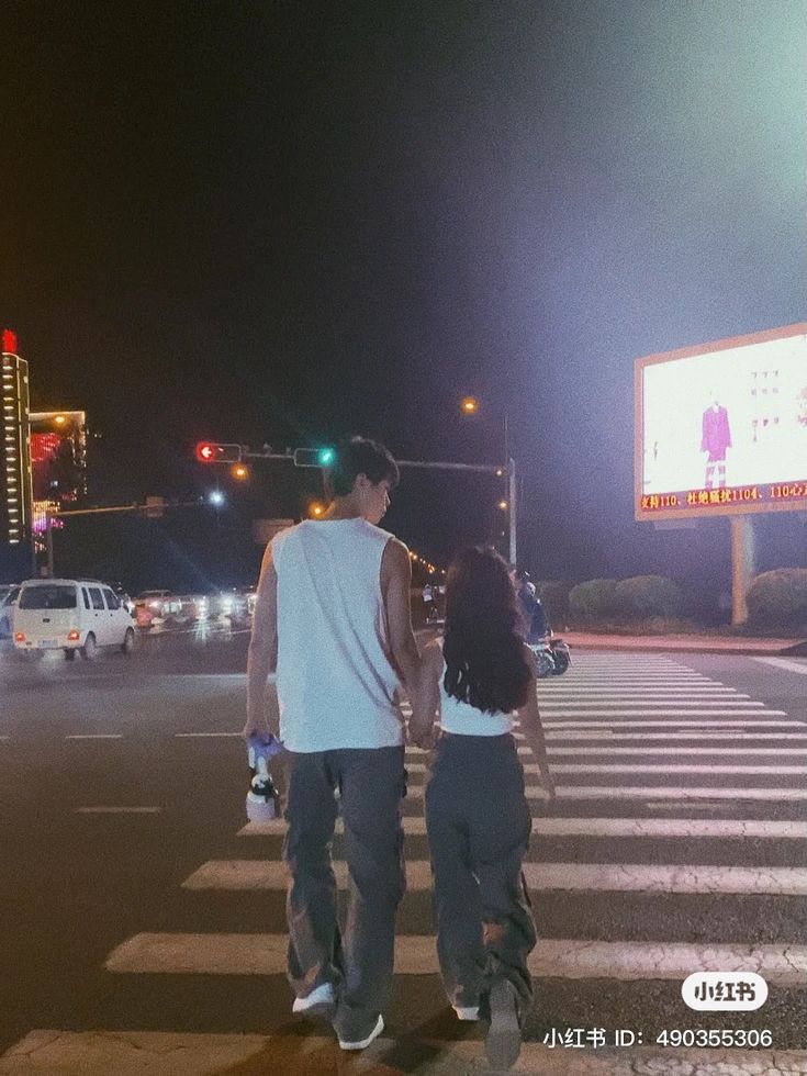 a man and woman walking across a cross walk at night with cars in the background