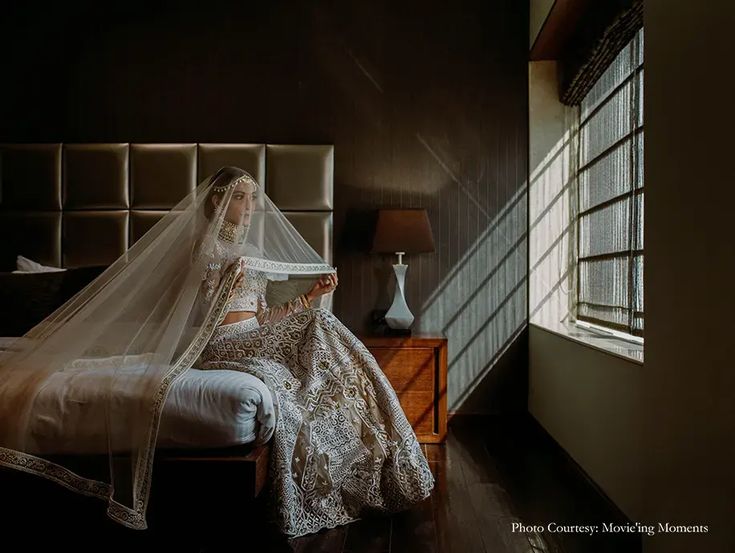 a woman sitting on top of a bed wearing a wedding dress and holding a veil