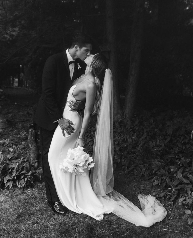 a bride and groom kissing in front of some trees at their wedding day, black and white photo