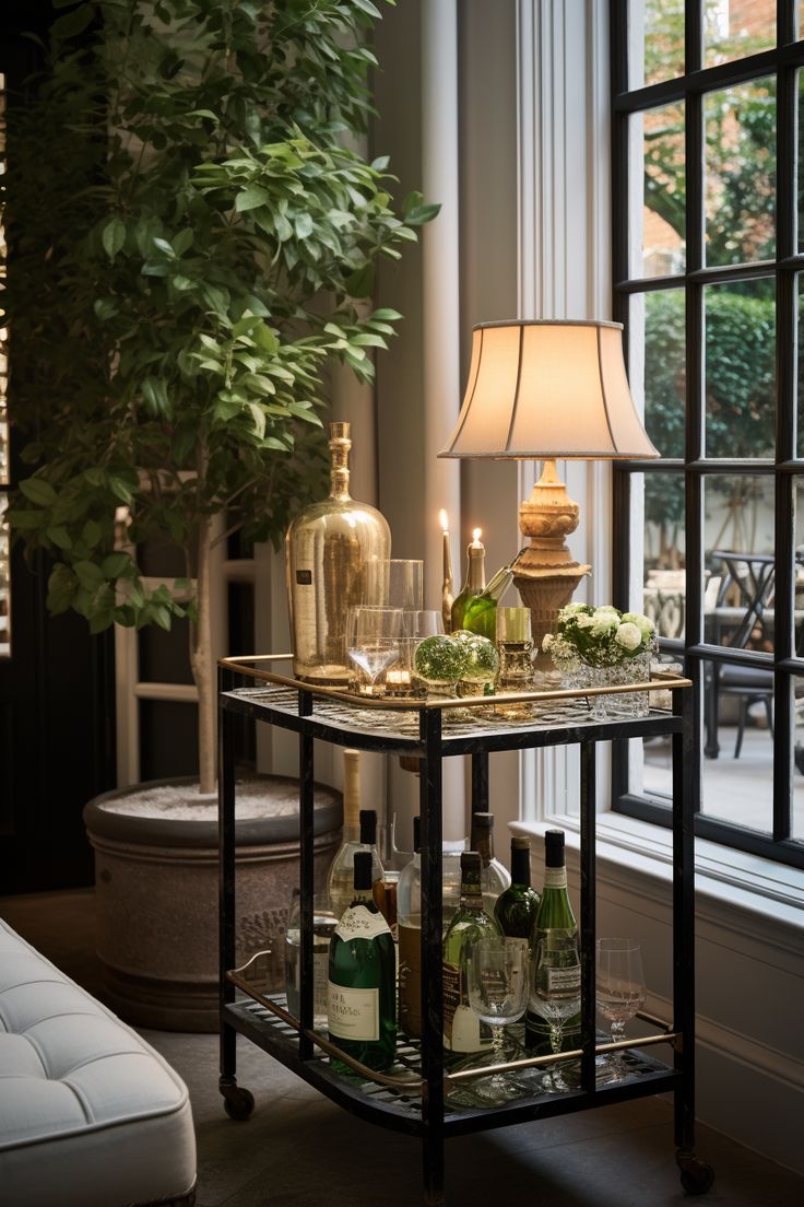 a bar cart with bottles and candles on it in front of a large window next to a couch