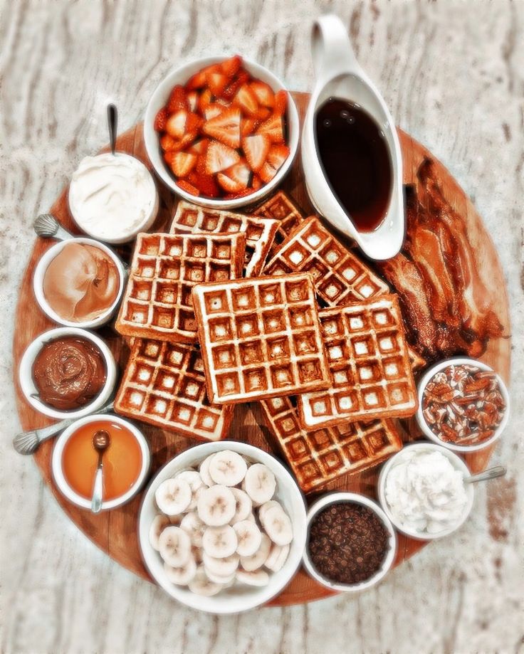 a wooden plate topped with waffles covered in syrup and toppings next to other foods