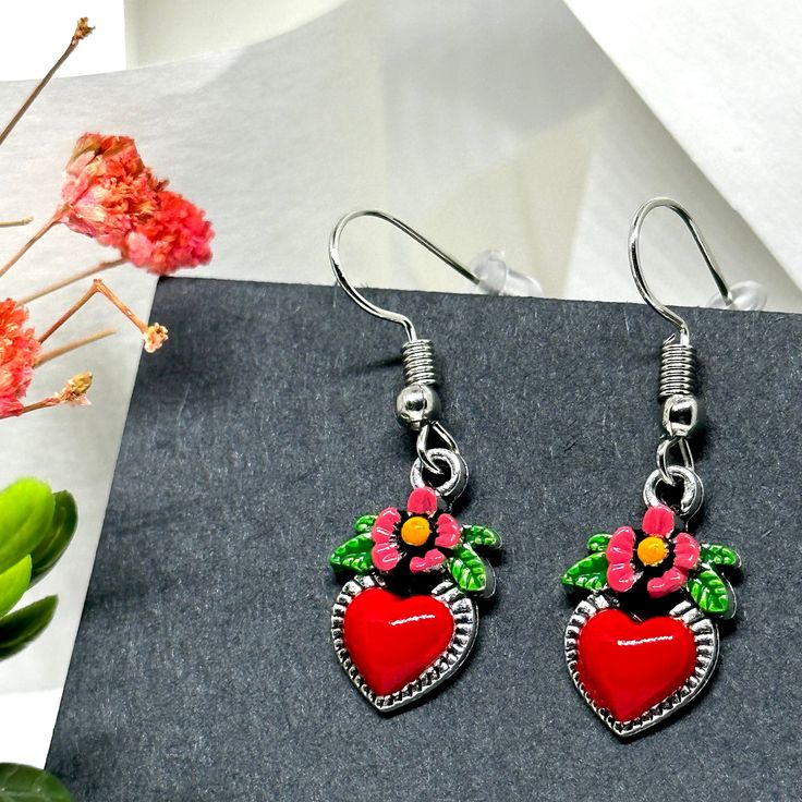 These Dainty Heart Earrings are the ultimate must-have. Expertly handcrafted and exquisitely hand-painted with a bold red enamel finish, playful pink flowers, and vibrant green leaves. Perfect for ladies and girls alike, these adorable dangling heart earrings are an ode to Frida's unique style. A perfect gift for any Fridalover, these Mexican-inspired accents are sure to put a smile on their face. Looking for a charming, one-of-a-kind present that won't break the bank? Look no further than these delightful hand-painted heart earrings inspired by Frida! Each earring is carefully crafted and meticulously painted by hand in our studio, and it takes a considerable amount of time and extreme attention to detail to finish them and finally offer them for sale. “It is always the simple that produc Valentine's Day Heart Charm Enamel Earrings, Valentine's Day Enamel Dangle Earrings, Red Dangle Flower Earrings For Valentine's Day, Enamel Earrings With Heart Charm As Gift, Enamel Heart Charm Earrings As Gift, Pink Hand Painted Heart Jewelry, Cute Red Flower Jewelry, Hand Painted Heart Earrings For Valentine's Day Gift, Valentine's Day Heart-shaped Flower Earrings For Pierced Ears