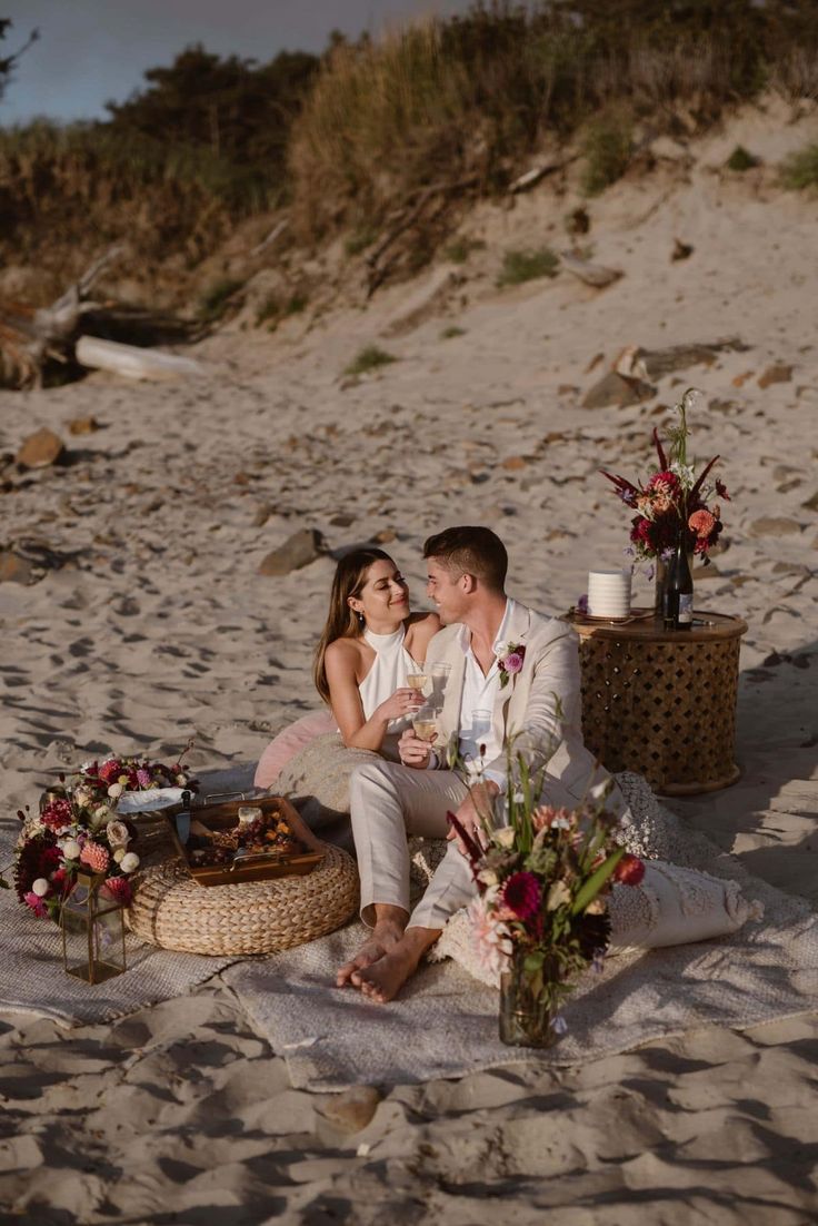 a man and woman sitting on the beach next to each other with flowers in front of them