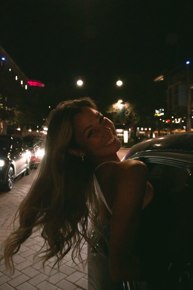 a woman standing next to a car on a street at night with her hair blowing in the wind