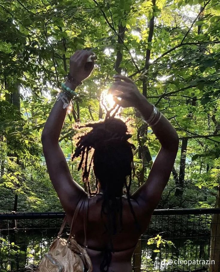 a woman with dreadlocks standing in front of trees and holding her hands up to the sky