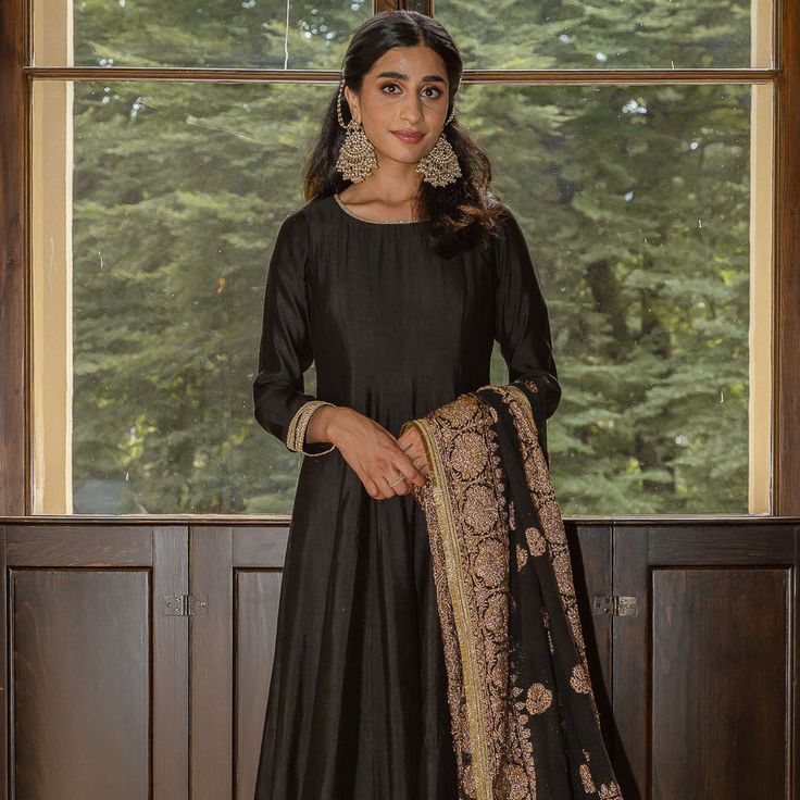 a woman standing in front of a window wearing a black dress with gold details on it