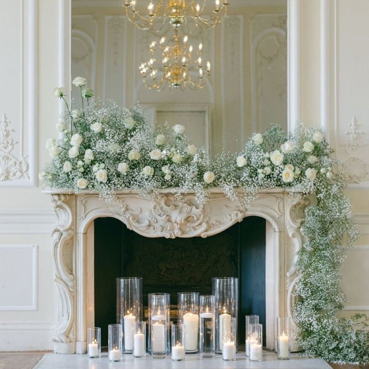 a fireplace with candles and flowers on it in front of a chandelier filled with greenery