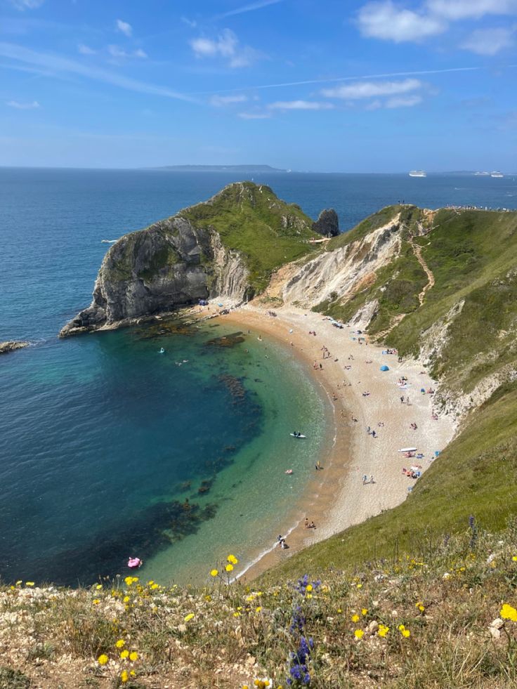 people are on the beach and in the water near an island with many rocks, grass and flowers