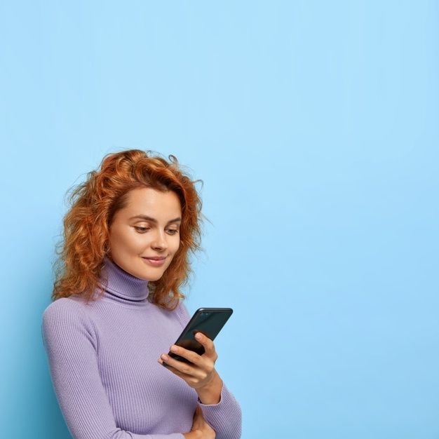 a woman with red hair is looking at her cell phone while standing against a blue background