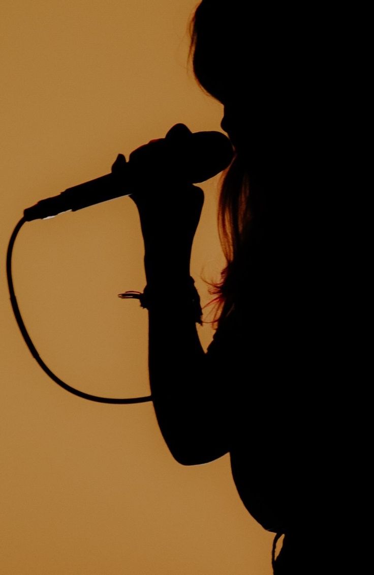 the silhouette of a woman holding a microphone in front of an orange sky with clouds