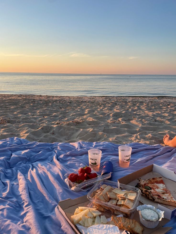 a woman sitting on top of a blanket next to a box of food at the beach