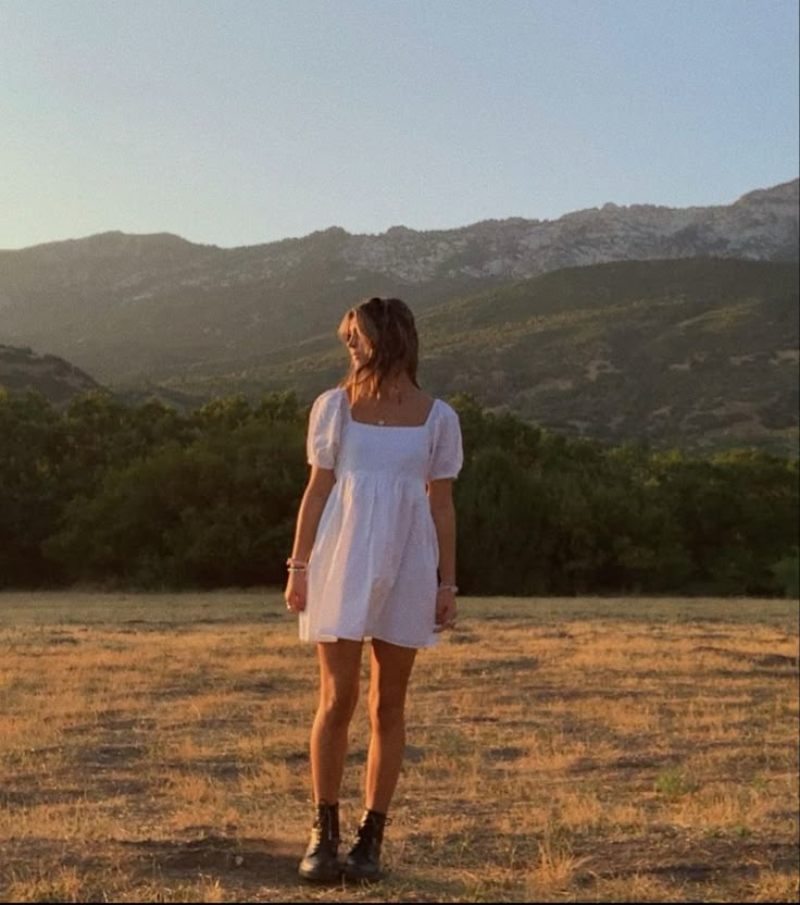 a woman in a white dress standing on top of a dry grass field with mountains in the background