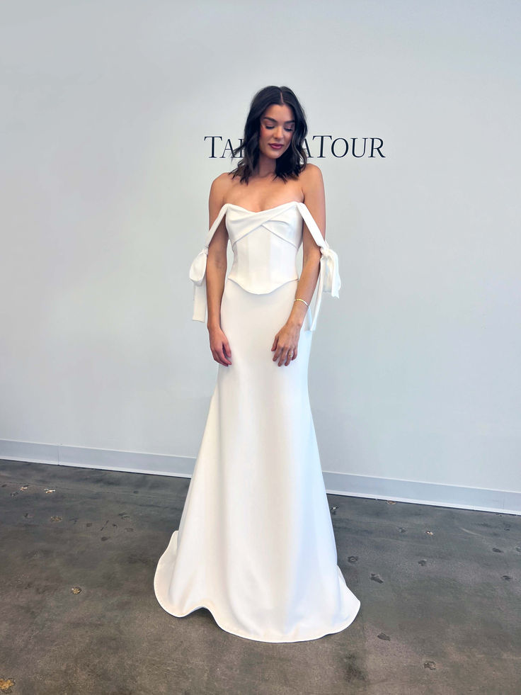 a woman standing in front of a wall wearing a white dress with an off the shoulder bow