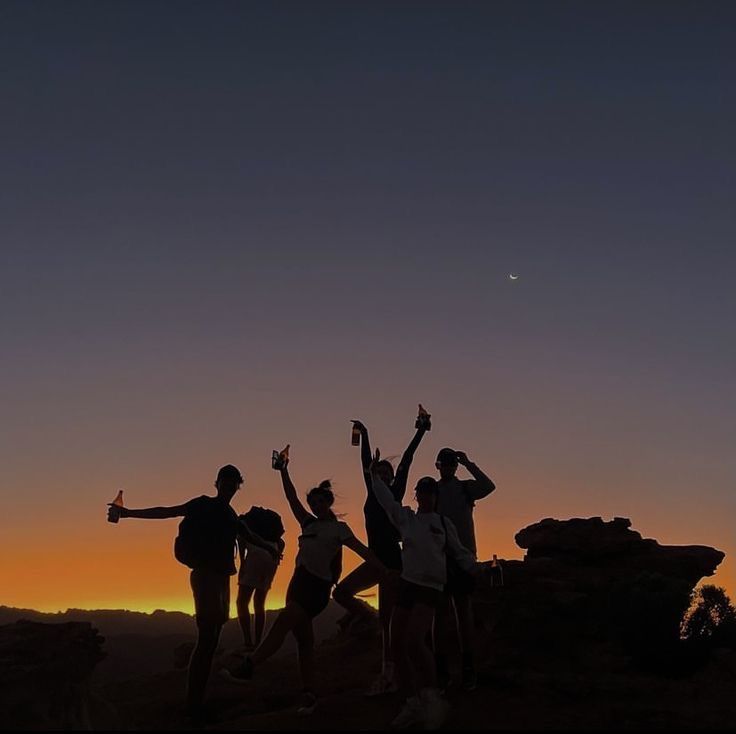 four people standing on top of a hill with their arms in the air at sunset