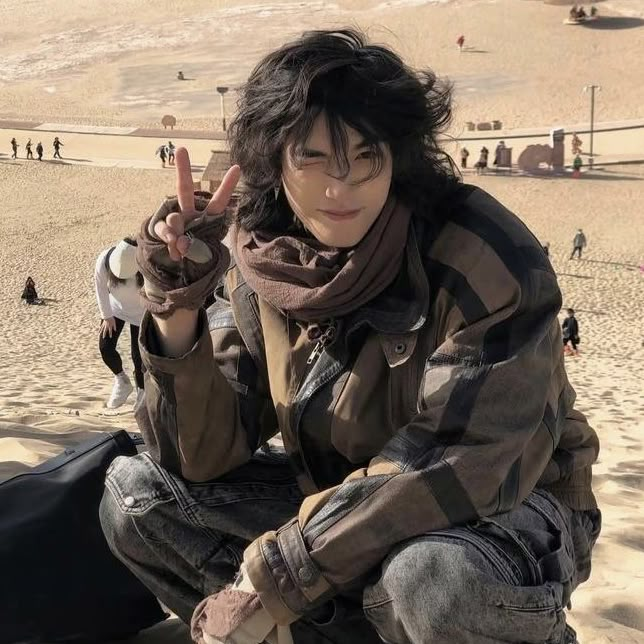 a man sitting on top of a sandy beach next to a black bag holding a peace sign