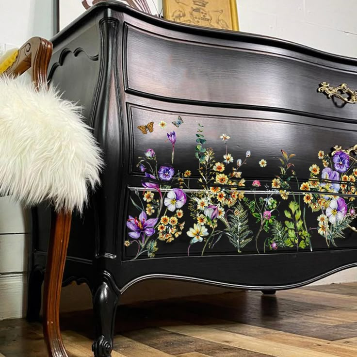 a black dresser with flowers painted on the front and sides, next to a chair