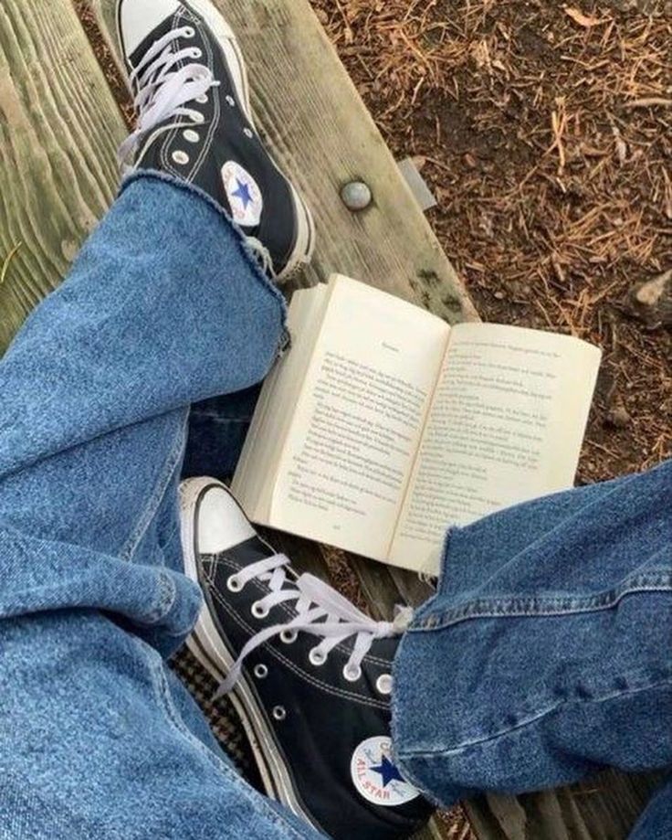 someone is reading a book while sitting on a bench with their legs crossed and wearing converse shoes