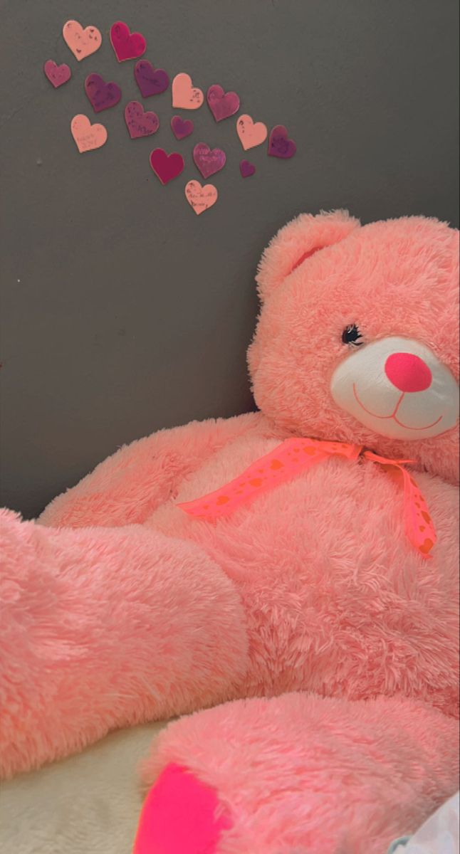 a large pink teddy bear sitting next to a wall with hearts on it's back