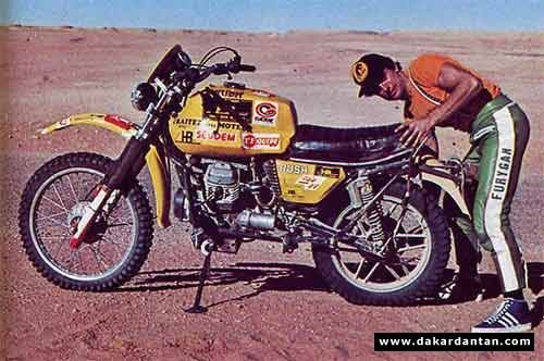 a man leaning on a yellow and black motorcycle in the middle of desert with his hand on the seat