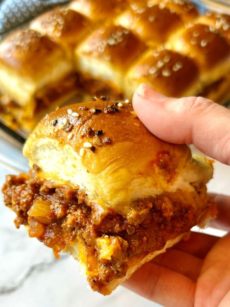 a hand holding up a piece of bread with meat and cheese on it in front of a pan