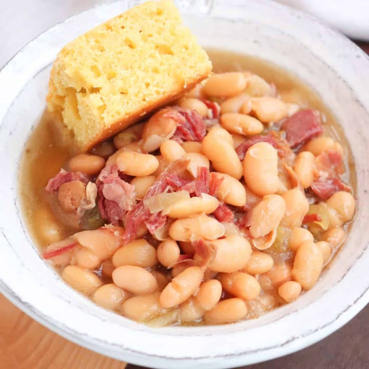 a white bowl filled with beans and ham next to a piece of bread on top of a wooden table