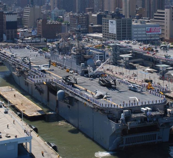 an aircraft carrier is docked in the water with other boats and large buildings behind it