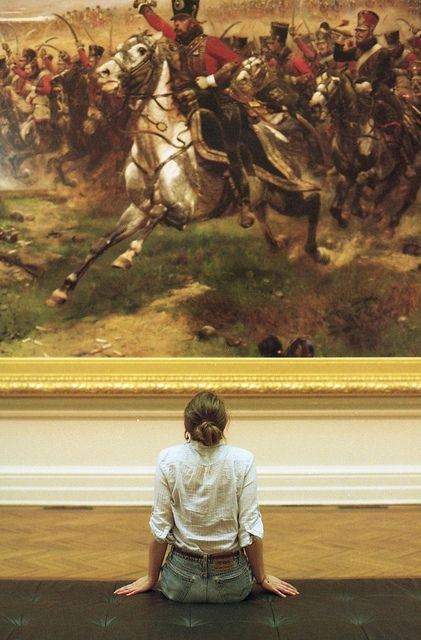 a woman sitting on the ground in front of a painting with people on horses behind her