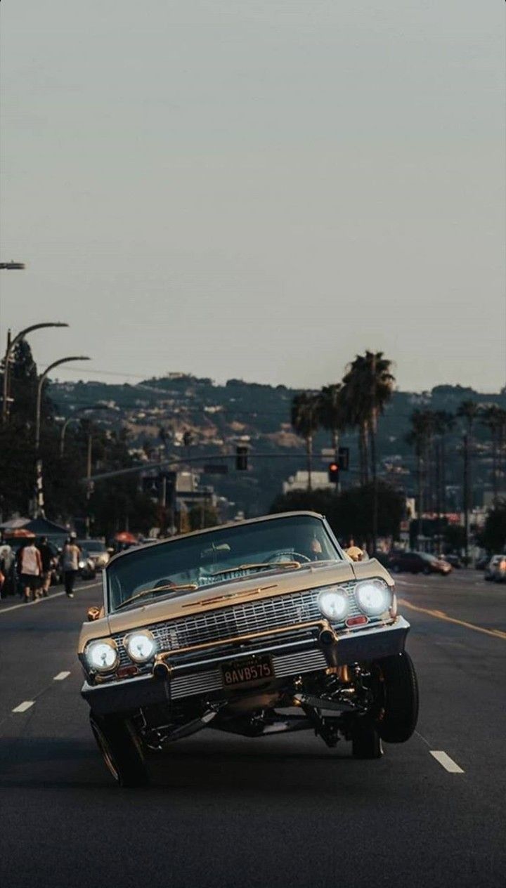 an old car driving down the street in front of some people and traffic lights with palm trees behind it