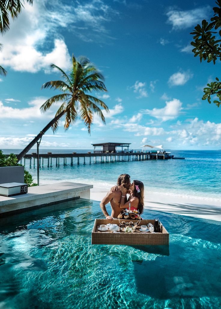 two people are sitting in a boat on the water with palm trees and a pier in the background