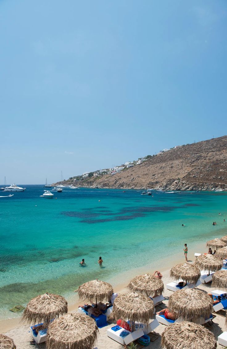 many umbrellas and chairs are on the beach near the blue water with boats in the background