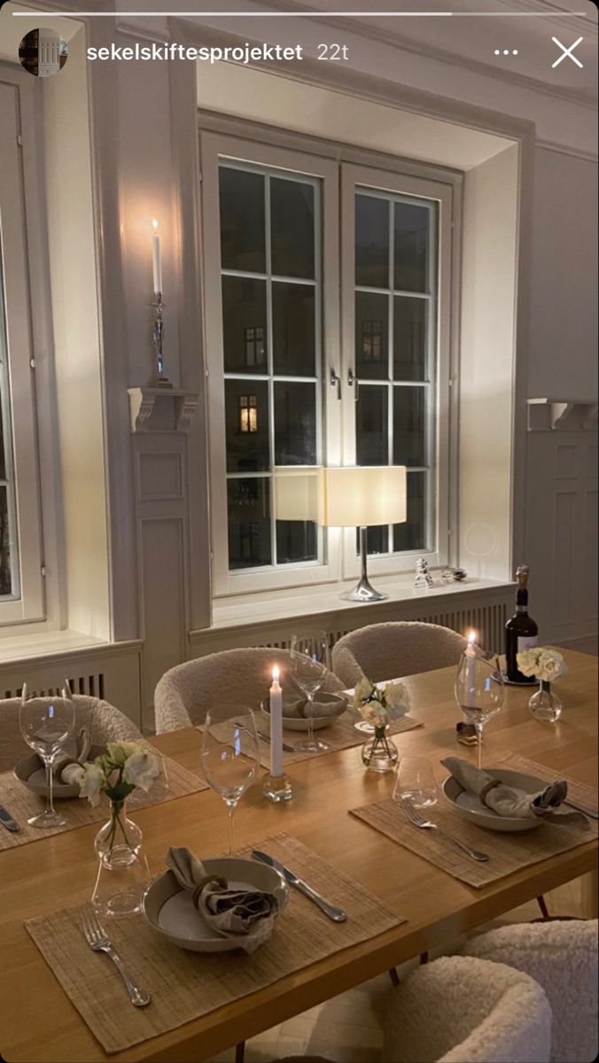 a dining room table set with place settings and candles on the dinner tables in front of two windows