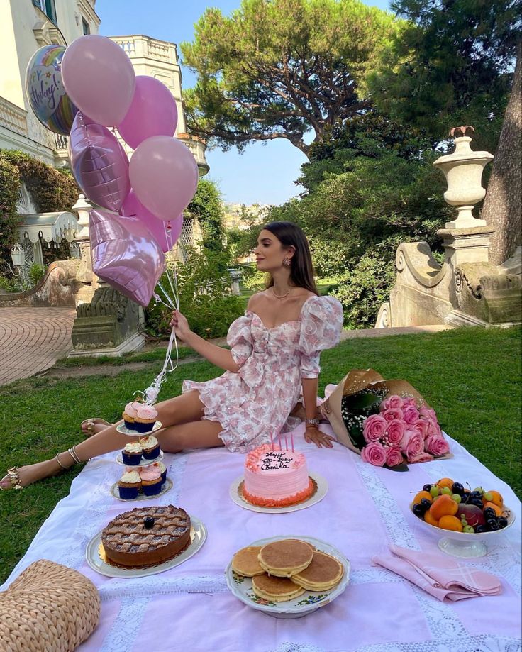 a woman sitting on the grass with some cake and balloons in front of her at a picnic