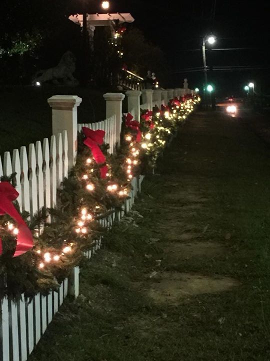 a white picket fence covered in christmas lights and wreaths with red bows on them