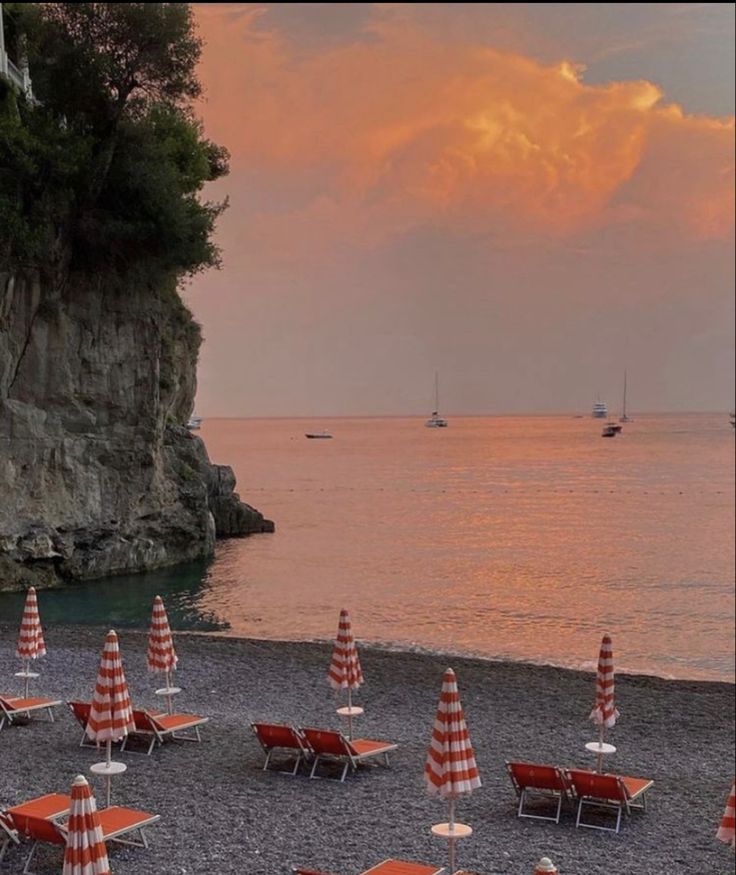 beach chairs and umbrellas are on the sand near the water at sunset with boats in the distance