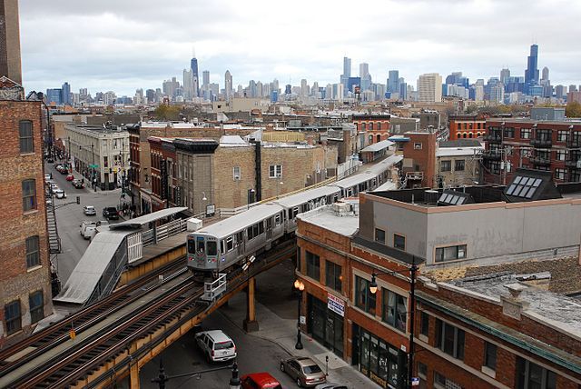 a train traveling through a city with tall buildings in the backgroung and cars parked on the street