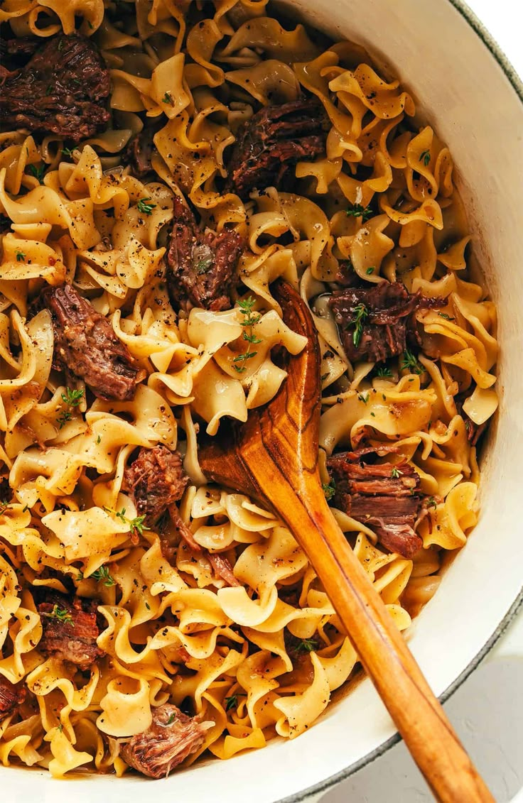 a pot filled with beef and noodles next to a wooden spoon