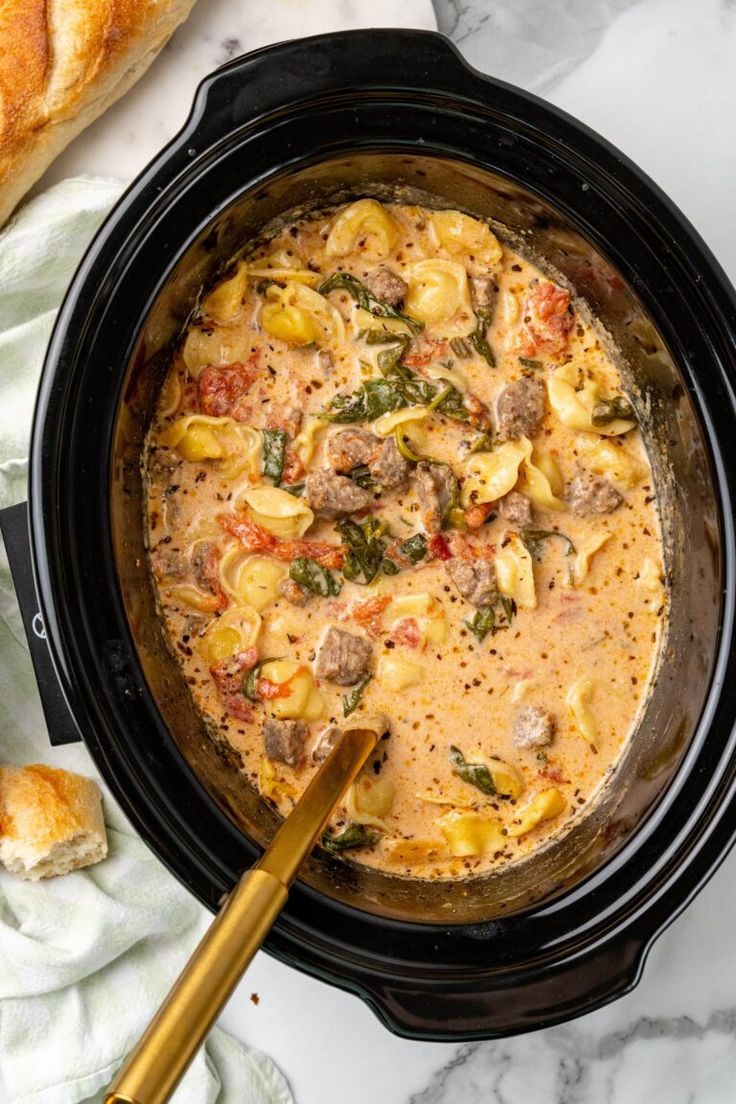 a crock pot filled with pasta, meat and spinach soup next to bread