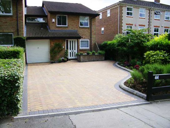 a brick driveway in front of two houses with bushes and shrubs on either side of the driveway