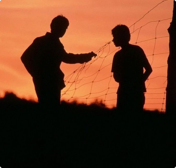 two men standing next to each other in front of a barbed wire fence at sunset