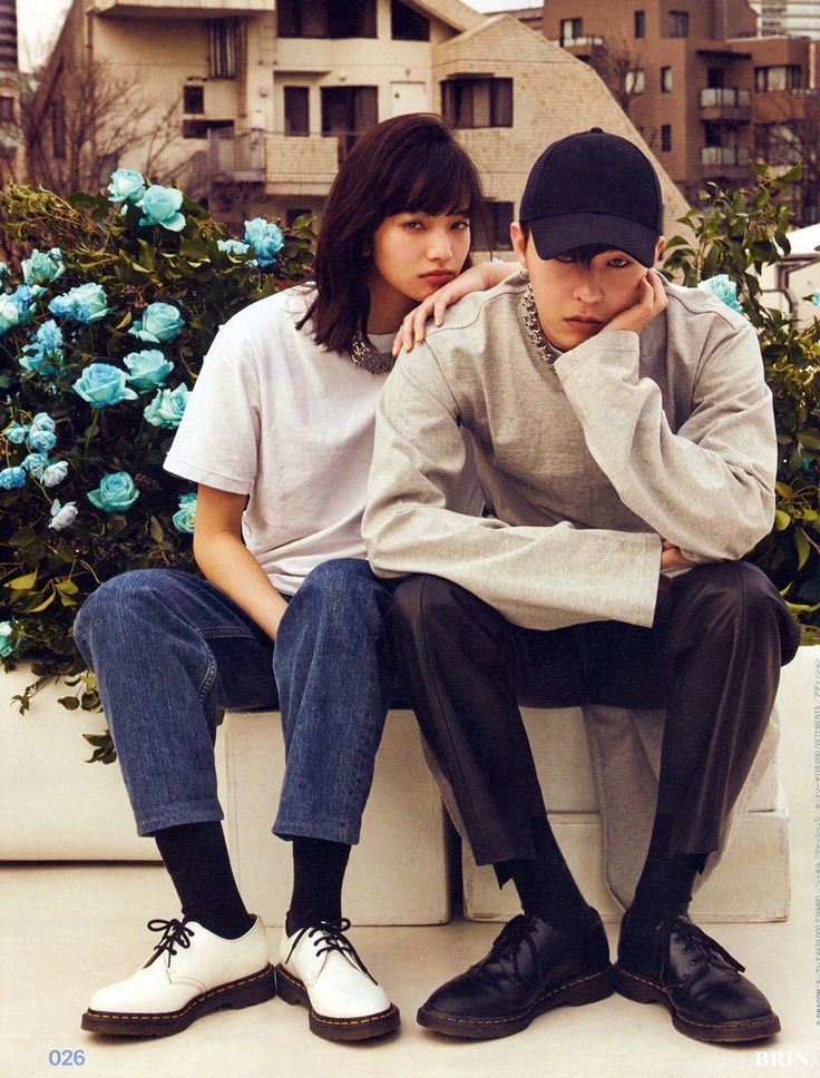 two people are sitting on a bench in front of blue flowers and building behind them