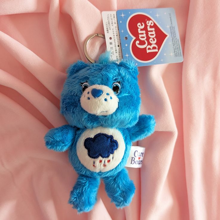 a blue teddy bear keychain is laying on a pink sheet with a tag