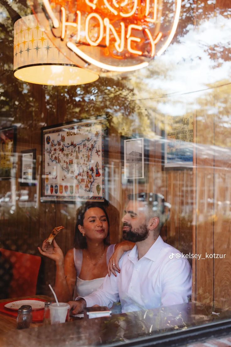 a man and woman sitting at a table in front of a restaurant window eating pizza