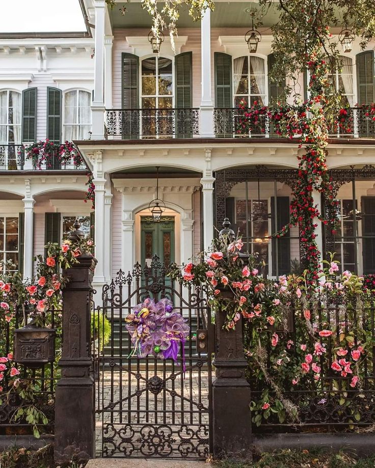 an iron gate with flowers growing on it in front of a white house that has many windows and balconies