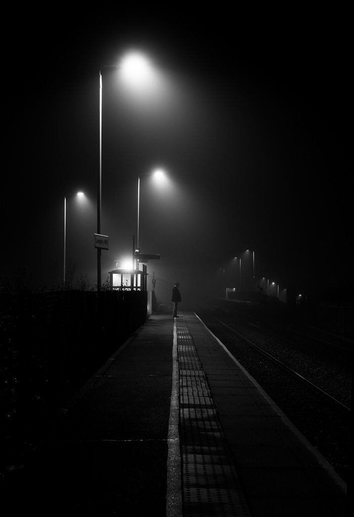 black and white photograph of person walking on train tracks at night with street lights in fog