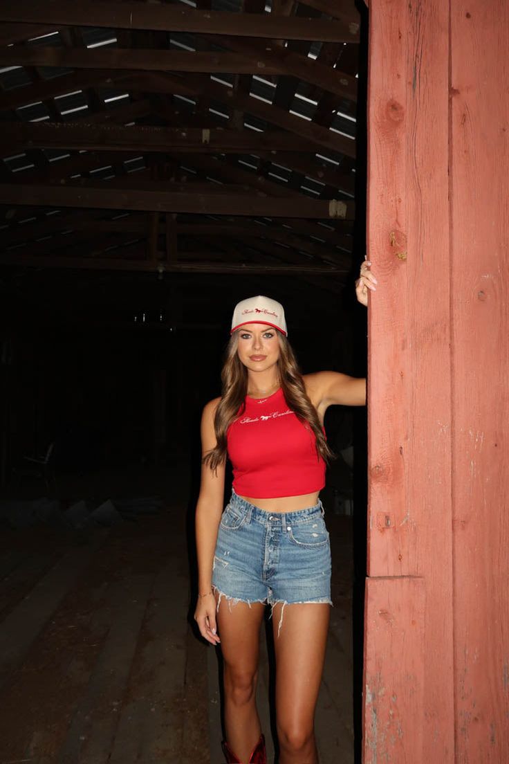 a woman in red shirt and denim shorts standing next to a wooden structure with her hand on the wall