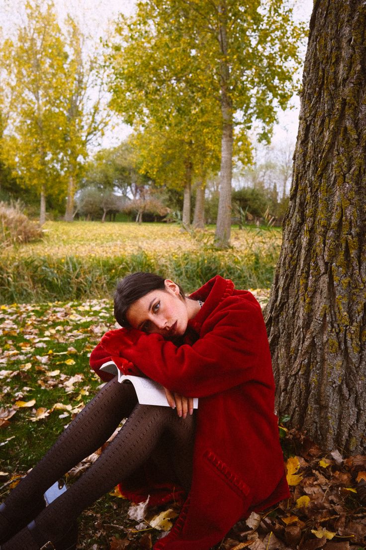 a woman in a red coat sitting next to a tree with her hands on her head