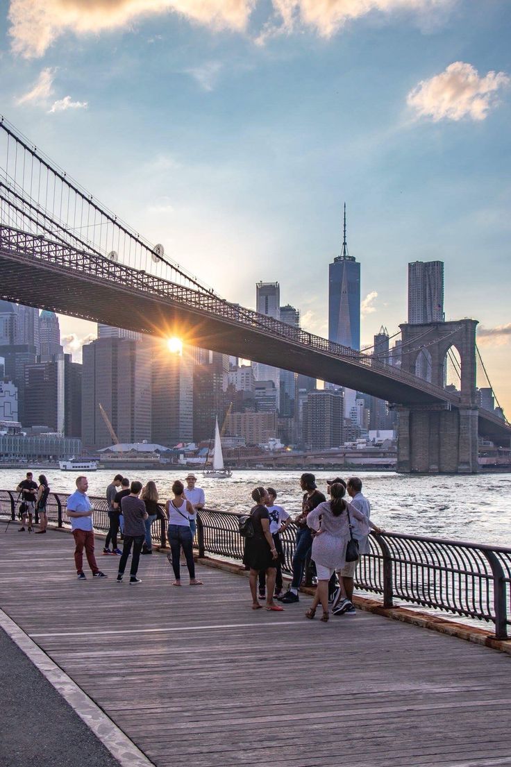 many people are walking on the boardwalk by the water and bridge in new york city