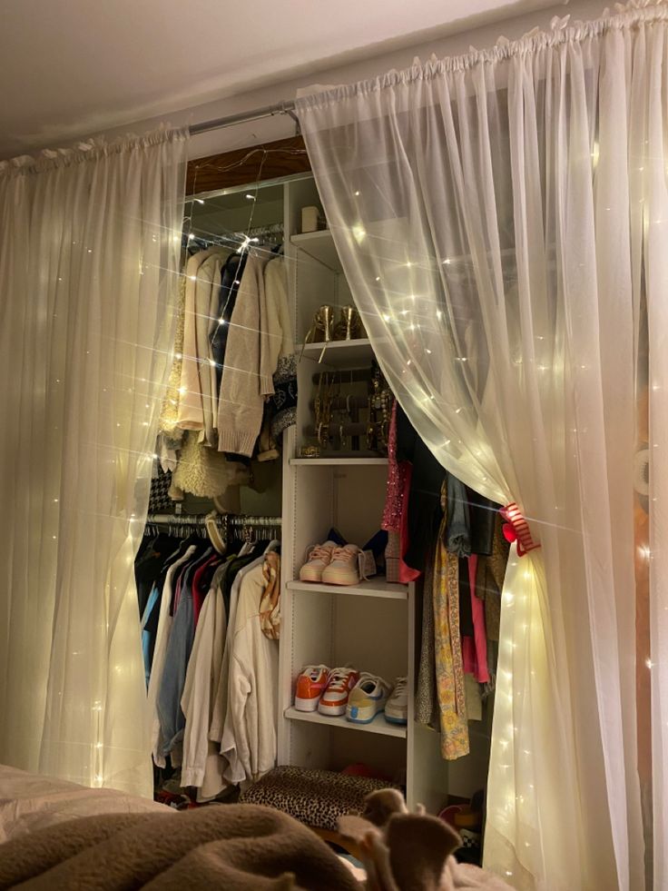 an open closet with clothes and shoes hanging on the shelves next to a curtained window