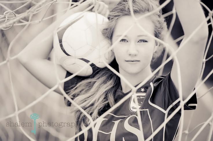 a young woman holding a soccer ball behind a goalie net with her hair blowing in the wind