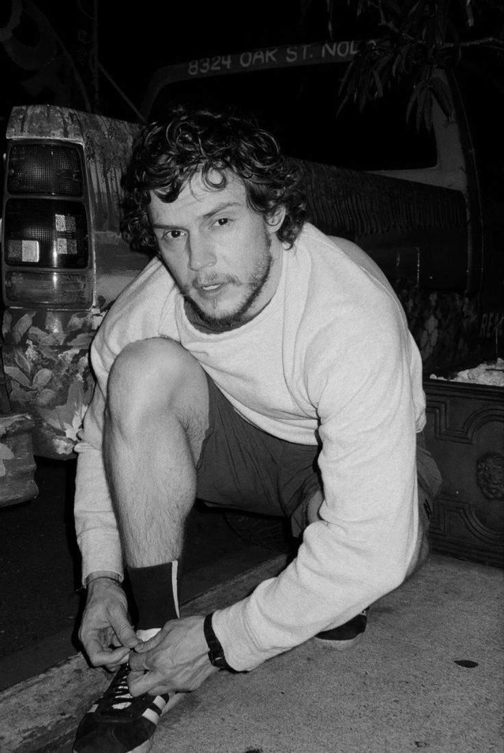 black and white photograph of a man tying his shoes on the ground in front of a van